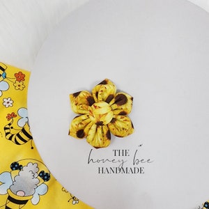 May include: A yellow and brown sunflower fabric flower with a white background. The text "THE honey bee HANDMADE" is written below the flower.