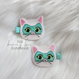 May include: Two white glitter cat hair clips with pink ears and green eye masks. The text "THE honey bee HANDMADE" is visible below the clips.