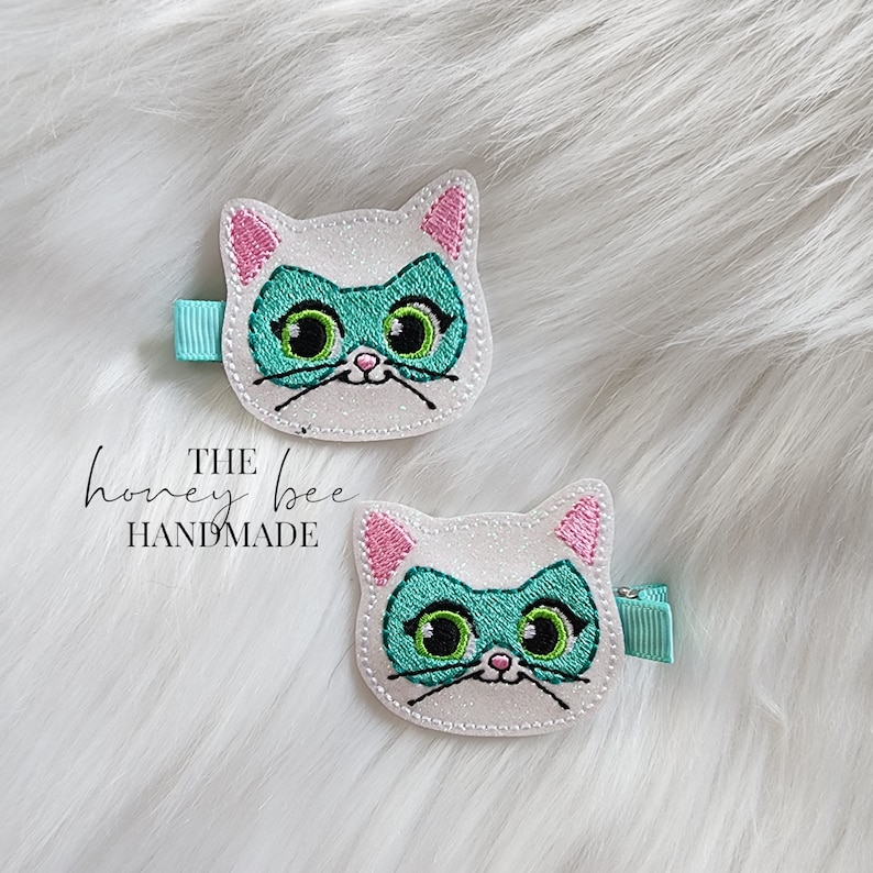 May include: Two white glitter cat hair clips with pink ears and green eye masks. The text "THE honey bee HANDMADE" is visible below the clips.