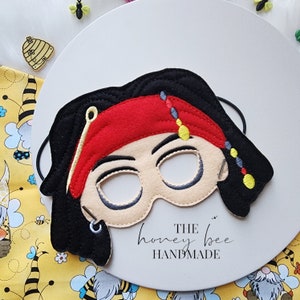 May include: Felt pirate eye mask with black hair, a red bandana, and gold and black beads. The mask has a stitched-on eye patch and a gold earring. The mask is handmade by The Honey Bee.