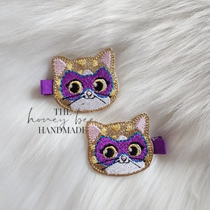 May include: Two gold glitter cat hair clips with purple masks and white eyes. The text "THE honey bee HANDMADE" is visible below the clips.