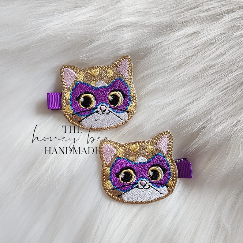 May include: Two gold glitter cat hair clips with purple masks and white eyes. The text "THE honey bee HANDMADE" is visible below the clips.
