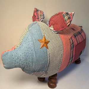 May include: A stuffed pig figurine crafted from various fabric patches including blue denim, houndstooth, and red plaid. The pig features a small metallic star on its side, pointed ears, and four dark brown wooden ball-shaped legs for support.