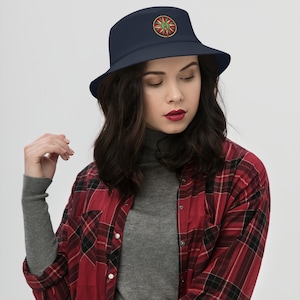 May include: A navy blue bucket hat featuring a circular compass rose emblem on the front. The emblem displays a detailed star design in shades of red, green, and gold, centered on the crown of the hat. The hat has a wide, downward-sloping brim.