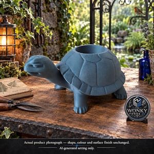 May include: A matte blue turtle-shaped planter with a textured shell and a circular opening on the back. The turtle has a smooth head and four legs, positioned on a wooden surface next to a small circular sign that reads The Wonky Forge 3D.