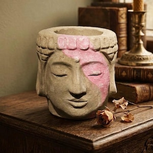 Könnte beinhalten: Ein beigefarbener, steinartiger Übertopf in Form eines menschlichen Kopfes mit geschlossenen Augen und strukturierter Oberfläche. Ein Teil der Stirn und der linke Augenbereich sind mit mattem rosa Pigment bemalt. Der Kopf hat eine obere Öffnung.
