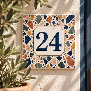 Puede incluir: Una placa cuadrada de número de casa con el número 24 en azul oscuro, centrado sobre un fondo blanco con un borde dorado. El borde está decorado con un patrón de terrazo colorido que consiste en fragmentos de piedra azules, verdes, naranjas y rosas.