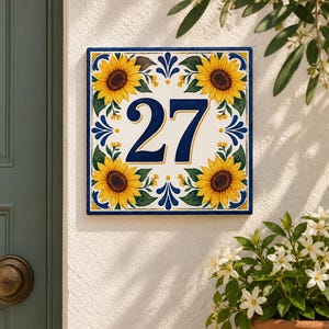 Op de afbeelding: Een vierkant keramieken huisnummerbordje met het getal 27 in donkerblauw. Het ontwerp bevat vier gele zonnebloemen met groene bladeren in de hoeken, geaccentueerd door blauwe decoratieve krullen op een witte achtergrond met een blauwe rand.