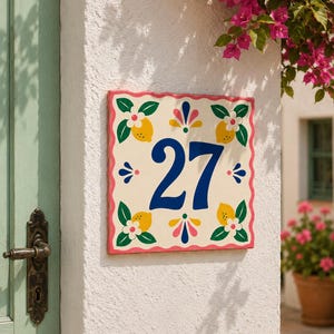 Op de afbeelding: Een vierkant keramieken huisnummerbordje met het getal 27 in een vetgedrukt blauw lettertype. Het ontwerp bevat gele citroenen met groene bladeren en witte bloemen in de hoeken, geaccentueerd door kleine roze en blauwe bloemmotieven.