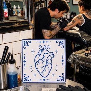 Può includere: Una piastrella quadrata bianca con un'illustrazione a linee blu di un cuore anatomico centrato all'interno di un bordo decorativo di stelle e accenti geometrici. La piastrella è posizionata su una superficie metallica vicino a attrezzature per tatuaggi.