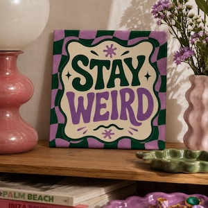 May include: A square canvas print featuring a cream-colored center with the dark green and purple text STAY WEIRD, surrounded by a wavy purple and green checkerboard border. The design includes small star and squiggle accents on the cream background.