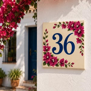 Puede incluir: Una placa de número de casa cuadrada de cerámica con el número 36 en una fuente serif azul oscuro. La placa tiene un fondo de color crema y está decorada con flores de buganvilla rosas pintadas y hojas verdes en las esquinas.