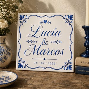 Może przedstawiać: Kwadratowa biała płytka ceramiczna z niebieską ozdobną ramką i motywami kwiatowymi. Napis "Lucía & Marcos" wykonano niebieską czcionką, nad nim znajduje się małe niebieskie serce, a na dole wydrukowano na niebiesko datę "18 . 07 . 2026".