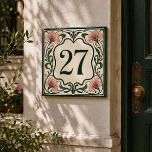 Może przedstawiać: Kwadratowa ceramiczna tabliczka z numerem domu przedstawiająca liczbę 27 ciemnozieloną czcionką szeryfową. Tabliczka ma kremowy środek z ozdobną obwódką z ciemnozielonych pnączy i czterema różowymi motywami kwiatowymi w rogach.