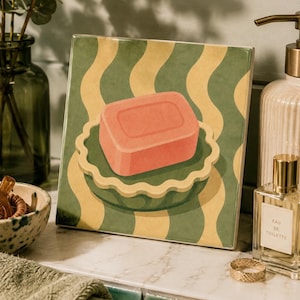 May include: A square ceramic tile featuring a graphic illustration of a rectangular pink soap bar resting in a scalloped green and yellow bowl. The background consists of vertical wavy stripes in alternating shades of muted green and pale yellow.