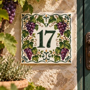 Puede incluir: Una placa de cerámica cuadrada para número de casa con el número 17 en verde oscuro. El diseño incluye racimos de uvas moradas pintados a mano, hojas de parra verdes y pequeñas flores blancas dispuestas en un borde simétrico sobre fondo crema.