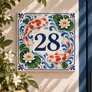 Könnte beinhalten: Ein quadratisches Hausnummernschild aus Keramik mit der Zahl 28 in Dunkelblau. Das Design zeigt zwei orange-weiße Koi-Fische, die im Kreis schwimmen, umgeben von weißen Seerosen, grünen Seerosenblättern und blauen, wirbelnden Wassermotiven.