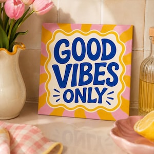 Può includere: Un cartello quadrato con il testo GOOD VIBES ONLY in grassetto blu. Lo sfondo presenta un motivo a raggi di sole ondulati gialli e rosa. Il cartello è incorniciato da un bordo bianco smerlato e si trova accanto a un vaso di tulipani rosa.