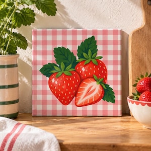 May include: A square wall art print featuring a vibrant illustration of two whole strawberries and one halved strawberry with green leaves, set against a light pink and white gingham checkered background pattern.