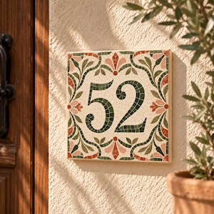 Op de afbeelding: Een vierkant huisnummerbordje met het getal 52 in donkergroene mozaïektegels. De rand is versierd met een symmetrisch patroon van groene bladeren en roze-oranje bloemmotieven op een crèmekleurige achtergrond.