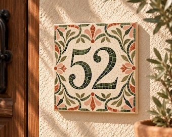 May include: A square house number plaque featuring the number 52 in dark green mosaic tiles. The border is decorated with a symmetrical pattern of green leaves and pinkish-orange floral motifs set against a cream-colored background.