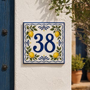 Puede incluir: Un azulejo cuadrado de cerámica para número de casa con el número 38 en azul oscuro. El diseño incluye un borde pintado a mano con limones amarillos, hojas verdes y flores blancas sobre un fondo crema, enmarcado por un patrón de líneas azules onduladas.