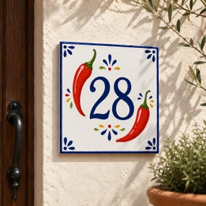 Könnte beinhalten: Ein quadratisches Hausnummernschild aus Keramik mit der Zahl 28 in Dunkelblau. Das Design zeigt zwei leuchtend rote Chilischoten, eine auf jeder Seite der Zahl, akzentuiert mit kleinen blauen, grünen und gelben Blumenmotiven und einem blauen Rand.