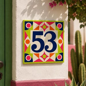 Könnte beinhalten: Ein quadratisches Hausnummernschild mit der Zahl 53 in Dunkelblau. Das Design zeigt ein weißes Zentrum, das von einem geometrischen Rand in Limettengrün, Pink, Orange und Dunkelblau umgeben ist und ein lebhaftes, farbenfrohes Dekor bildet.