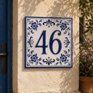 Könnte beinhalten: Ein quadratisches Hausnummernschild aus Keramik mit den Ziffern 46 in einer dunkelblauen Serifenschrift. Die weiße Fliese ist in jeder Ecke mit einem dunkelblauen Blumenmuster und einem dünnen blauen Rahmen verziert, montiert an einer beigen Wand.