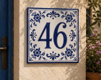May include: A square ceramic house number plaque featuring the digits 46 in a dark blue serif font. The white tile is decorated with a dark blue floral pattern in each corner and a thin blue border frame, mounted on a textured beige wall.