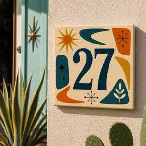 Könnte beinhalten: Ein quadratisches Hausnummernschild mit der Zahl 27 in Dunkelblau. Der cremefarbene Hintergrund zeigt bunte grafische Formen, darunter eine gelbe Sonne, orangefarbene und blaugrüne Bumerangs, einen Stern und ein blaugrünes Dreieck mit einem Blatt.