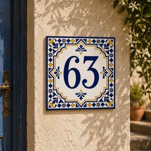 Op de afbeelding: Een vierkant keramisch huisnummerbordje met het getal 63 in donkerblauw. De tegel heeft een wit midden met een decoratieve rand in blauw, geel en wit, met geometrische patronen en bloemmotieven op een gestructureerd muuroppervlak.