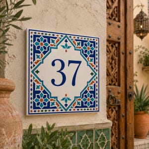 Könnte beinhalten: Ein quadratisches Hausnummernschild aus Keramik mit den Ziffern 37 in Dunkelblau. Das Design zeigt ein weißes Zentrum, umgeben von einer geometrischen Bordüre in Blau-, Türkis- und Orangetönen, auf einer strukturierten, cremeweißen Wand.