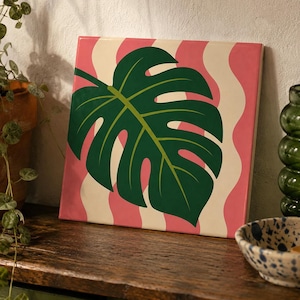 Può includere: Un quadro quadrato su tela raffigurante una grande illustrazione di una foglia di monstera verde scuro centrata su uno sfondo di strisce verticali ondulate in tonalità alternate di rosa tenue e crema, creando un design grafico audace.