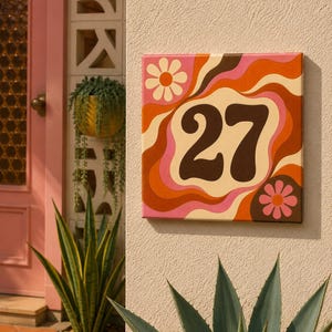 Könnte beinhalten: Ein quadratisches Hausnummernschild mit der Zahl 27 in Dunkelbraun vor einem Hintergrund aus wellenförmigen, geschwungenen Bändern in Orange-, Rosa- und Cremetönen, verziert mit zwei stilisierten Blumenmotiven in den gegenüberliegenden Ecken.