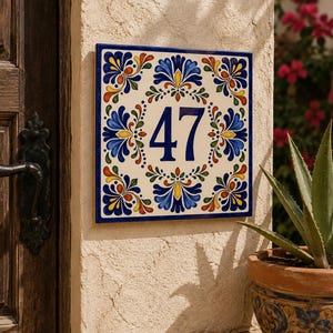 Op de afbeelding: Een vierkant keramisch huisnummerbordje met het getal 47 in donkerblauw. De tegel heeft een crèmekleurige achtergrond versierd met een symmetrisch bloemmotief in tinten blauw, geel, groen en oranje, omlijst door een donkerblauwe rand.