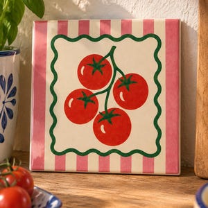 Può includere: Una piastrella quadrata in ceramica con un'illustrazione centrale di quattro pomodori rossi su un rametto verde. Il disegno è incorniciato da un bordo ondulato verde scuro e da un bordo a righe verticali rosa e bianche su fondo crema.