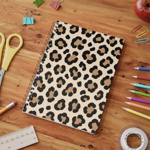 May include: A spiral-bound notebook featuring a repeating leopard print pattern with black spots on a tan background. The notebook is positioned on a wooden surface alongside various office supplies including scissors, coloured pencils, and a ruler.