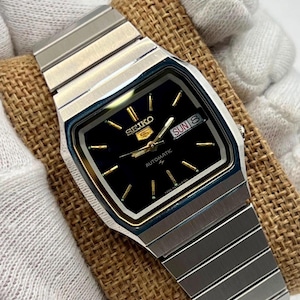 Puede incluir: Un reloj de pulsera automático Seiko 5 con esfera cuadrada negra, marcadores de hora y manecillas en tono dorado. El reloj incluye una ventana de día y fecha en la posición de las tres y una correa de eslabones de acero inoxidable plateado.