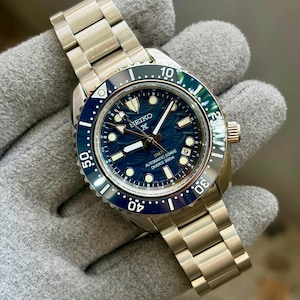 May include: A Seiko Prospex GMT automatic diver's watch with a stainless steel link bracelet. The watch features a deep blue dial with a wave pattern, a rotating blue and black bezel, luminous markers, and text reading Seiko, GMT, Automatic 3 Days, Diver's 300m.