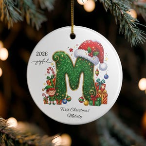 Puede incluir: Un adorno redondo de cerámica blanca con una letra M grande de color verde brillante que lleva un gorro de Papá Noel. El diseño incluye un pequeño elfo, cajas de regalo y bastones de caramelo. El texto dice 2026, joyful, First Christmas, Melody.