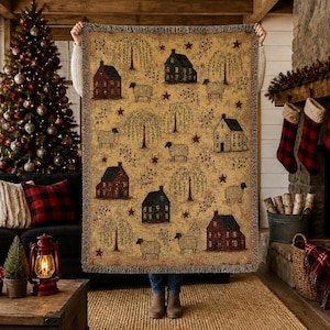 May include: A woven throw blanket featuring a repeating pattern of houses, sheep, willow trees, and stars in shades of tan, brown, black, and red. The rectangular textile has a fringed edge and displays a detailed folk-art style landscape scene.