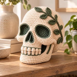 Puede incluir: Una calavera de ganchillo color crema con detalles en verde oscuro en las cuencas de los ojos, la cavidad nasal y la mandíbula. Una enredadera de hojas verde oscuro tejida a ganchillo está unida al lado de la calavera y los dientes son bolitas.