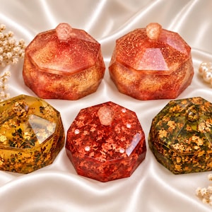 May include: Five octagonal trinket boxes with lids, featuring translucent resin bodies embedded with gold leaf flakes. Two boxes are deep red, two are amber-toned, and one is a vibrant red with small embedded crystals on the lid surface.