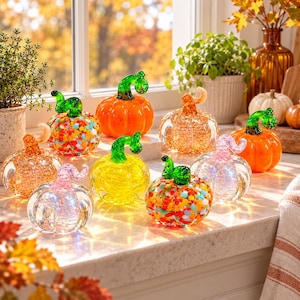 Op de afbeelding: Een verzameling van negen decoratieve glazen pompoenen in diverse kleuren en patronen, waaronder effen oranje, geel, transparant met glitters en veelkleurige confetti-ontwerpen. Elke pompoen heeft een opvallende, gebogen steel van groen of roze glas.