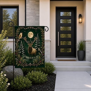 May include: A rectangular garden flag featuring a dark green background with a central illustration of a stag with large antlers standing in a snowy forest under a full moon. An owl and a small bird are perched on branches within a gold border.