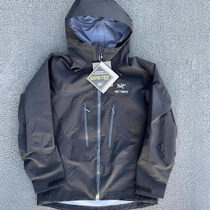 Può includere: Una giacca nera con cappuccio Arc'teryx dotata di cerniera frontale a tutta lunghezza, due tasche verticali sul petto con cerniere e un'etichetta Gore-Tex attaccata al colletto. La giacca presenta maniche lunghe con polsini regolabili e una fodera interna grigio chiaro.