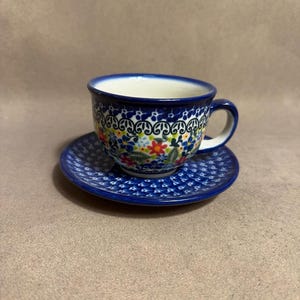 May include: A ceramic teacup and saucer set featuring a dark blue rim and a repeating pattern of small blue, red, and yellow flowers with green leaves. The cup has a curved handle and a white interior, while the saucer matches the floral design.