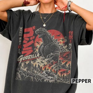 May include: A charcoal gray t-shirt featuring a large graphic print of Godzilla against a red sun. The design includes Japanese text, a city skyline, airplanes, and three smaller panels showing Godzilla's eye, silhouette, and a torii gate.