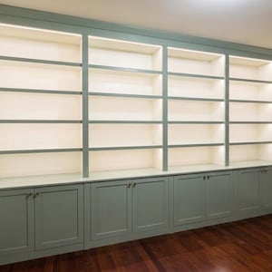 May include: A large, built-in wall unit featuring sage green cabinetry with lower storage cabinets and upper open shelving. The unit spans the width of the wall and includes integrated lighting within each shelf section, providing illumination.